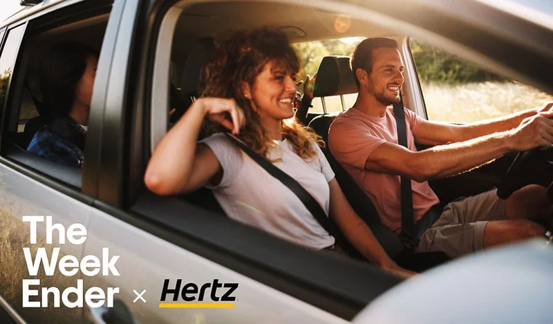 Image description: A man and a woman smile while driving in a car. The WeekEnder x Hertz