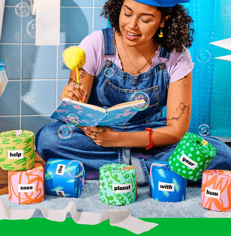 Image: Woman with a beret, a fluffy yellow pen, and a sky-themed journal, writing poetry, surrounded by the Poetry Edition of Who Gives A Crap toilet paper