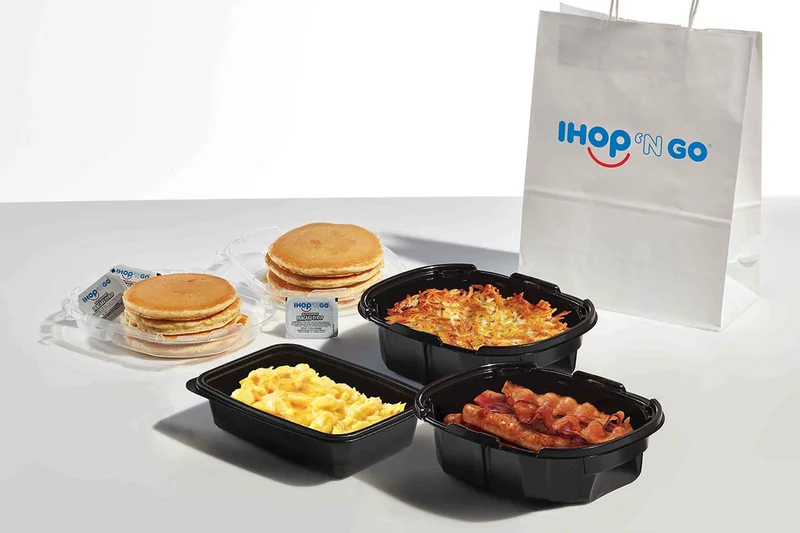 An assortment of food in catering trays, including pancakes, bacon and scrambled eggs, with a white IHOP N GO paper bag in the background.