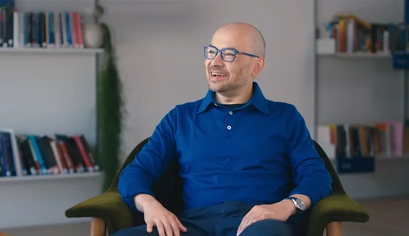 A man with glasses, wearing a blue collared shirt, sits in a green armchair and smiles while looking to his left. A bookshelf is visible in the background.