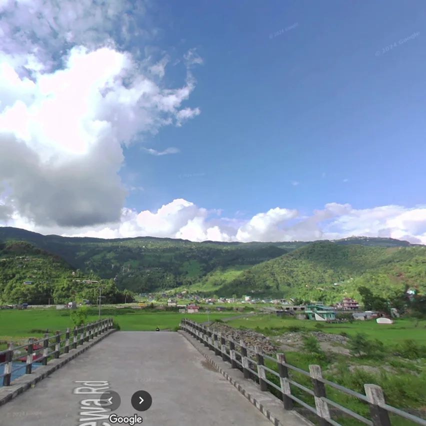 A Google Street View image shows a bridge crossing over a small river or stream, leading towards a village nestled in a valley. Lush green, forested hills rise on either side of the valley under a bright blue sky with scattered clouds.