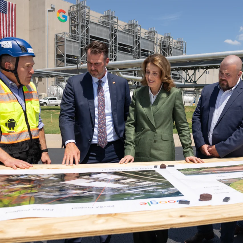 Four people, two in suits and two in work attire, stand around a large table with blueprints in front of a Google data center building.