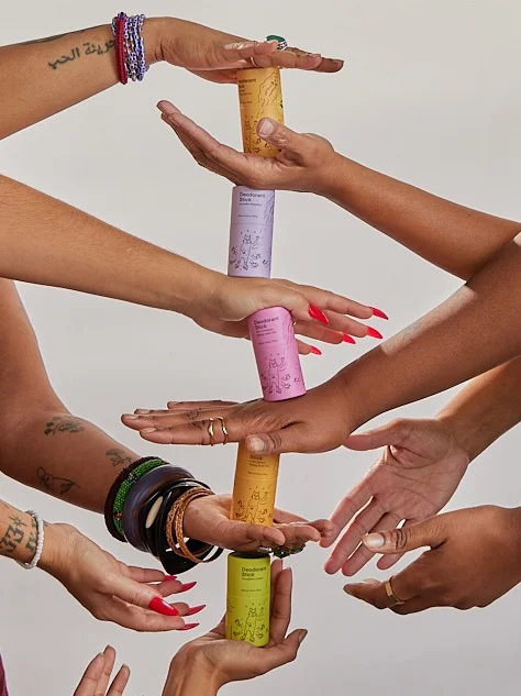 Many pairs of hands hold up colorful tubes of deodorant, all stacked atop each other.