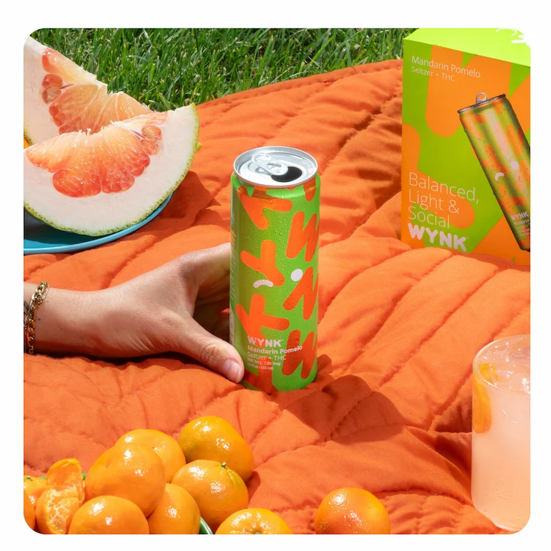 Photo of a can of Wynk Mandarin Pomelo on an orange picnic blanket on green grass, with mandarin oranges and a juicy pomelo on the blanket around.