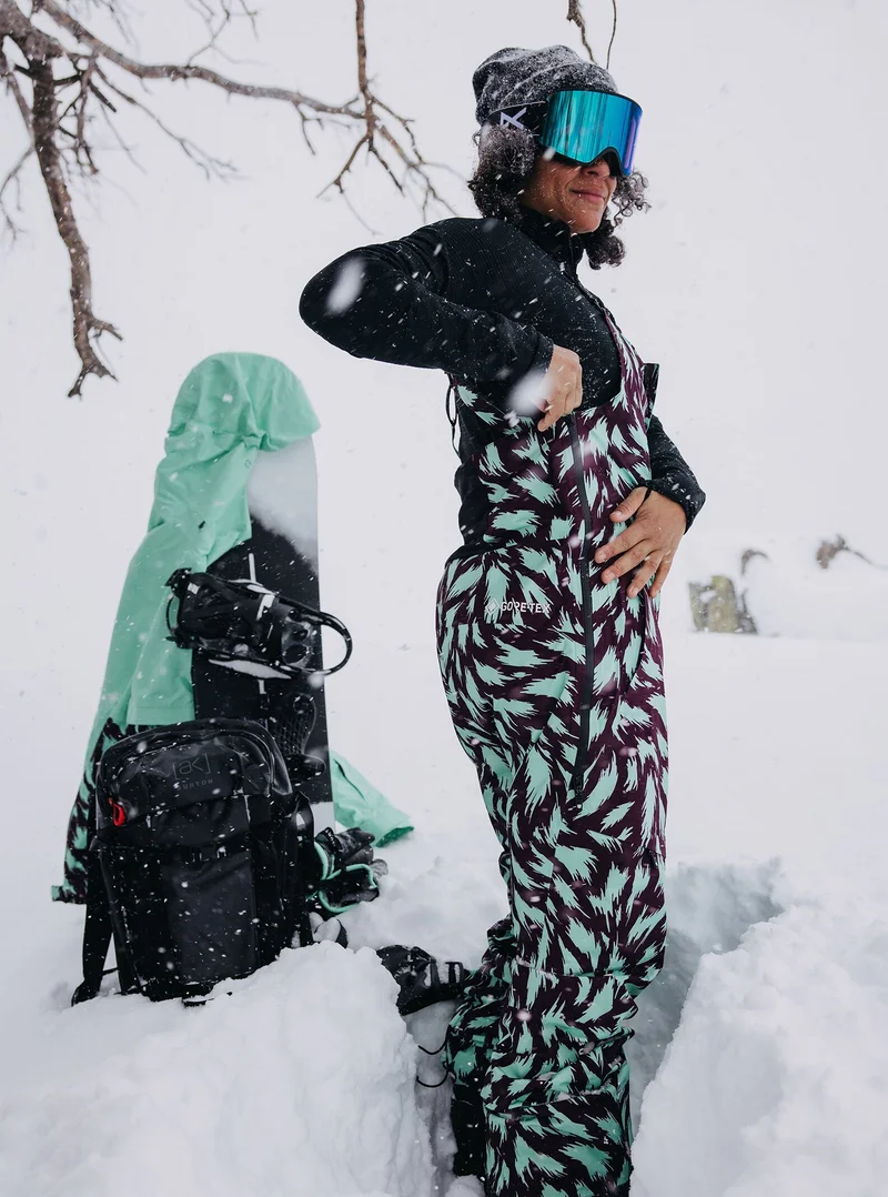 Women's Burton [ak] Kimmy GORE-TEX 2L Bib Pants in a teal and purple fur pattern on a model in the deeo snow with a Burton Jacket and Snowboard in the background