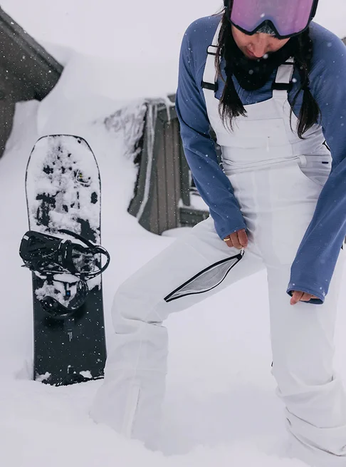 Women's Burton Avalon 2L Stretch Bib Pants in white on a model the snow with a Burton Snowboard in the background