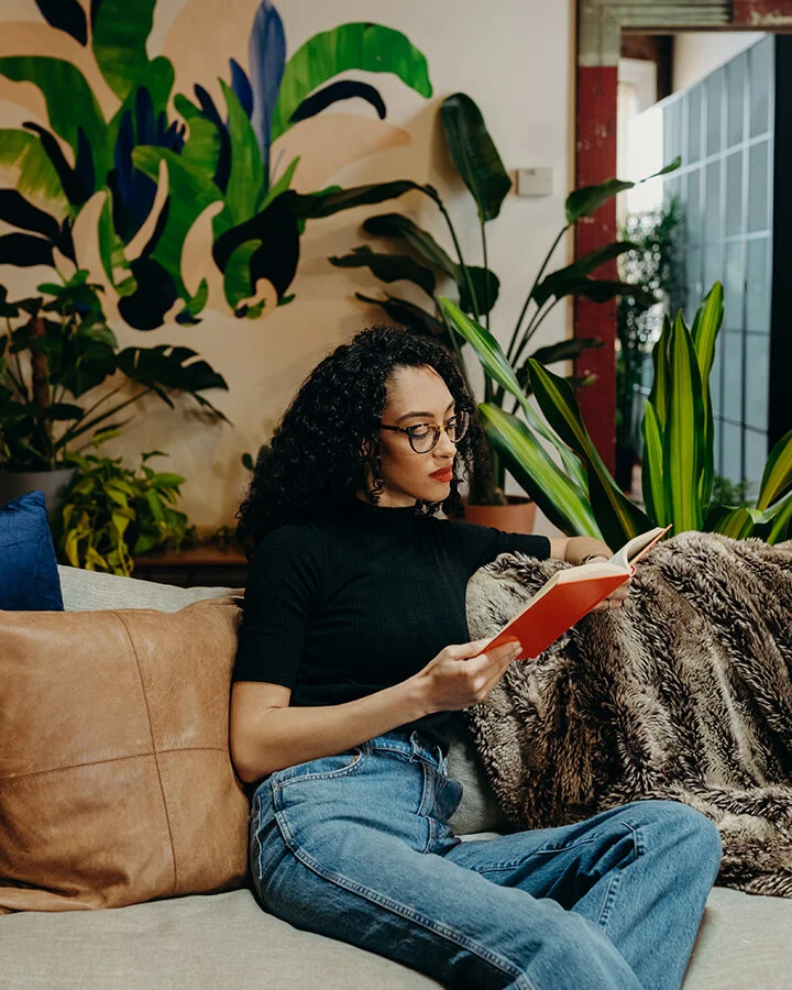 woman reading while sitting on couch surrounded by plants