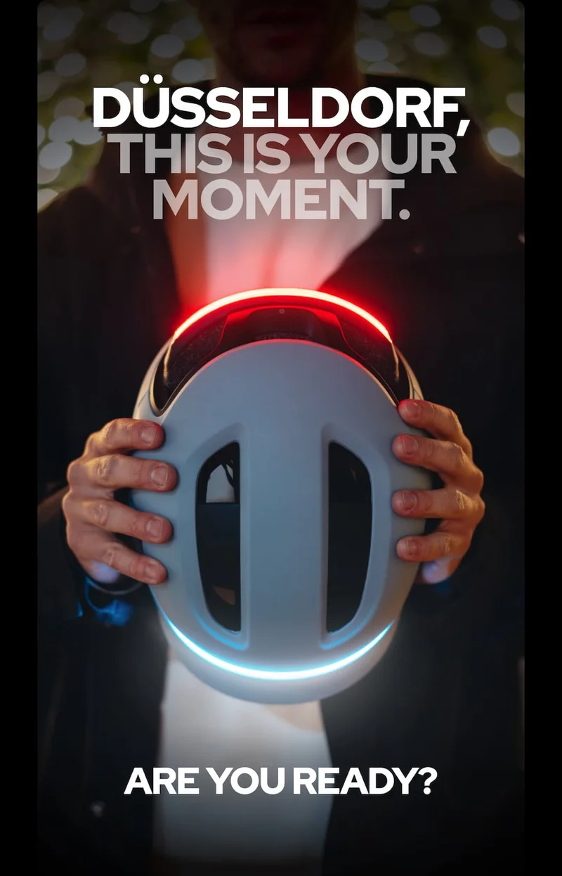 Person holding a UNIT 1 helmet with illuminated front and rear lights, with text reading “DÜSSELDORF, THIS IS YOUR MOMENT. ARE YOU READY?”
