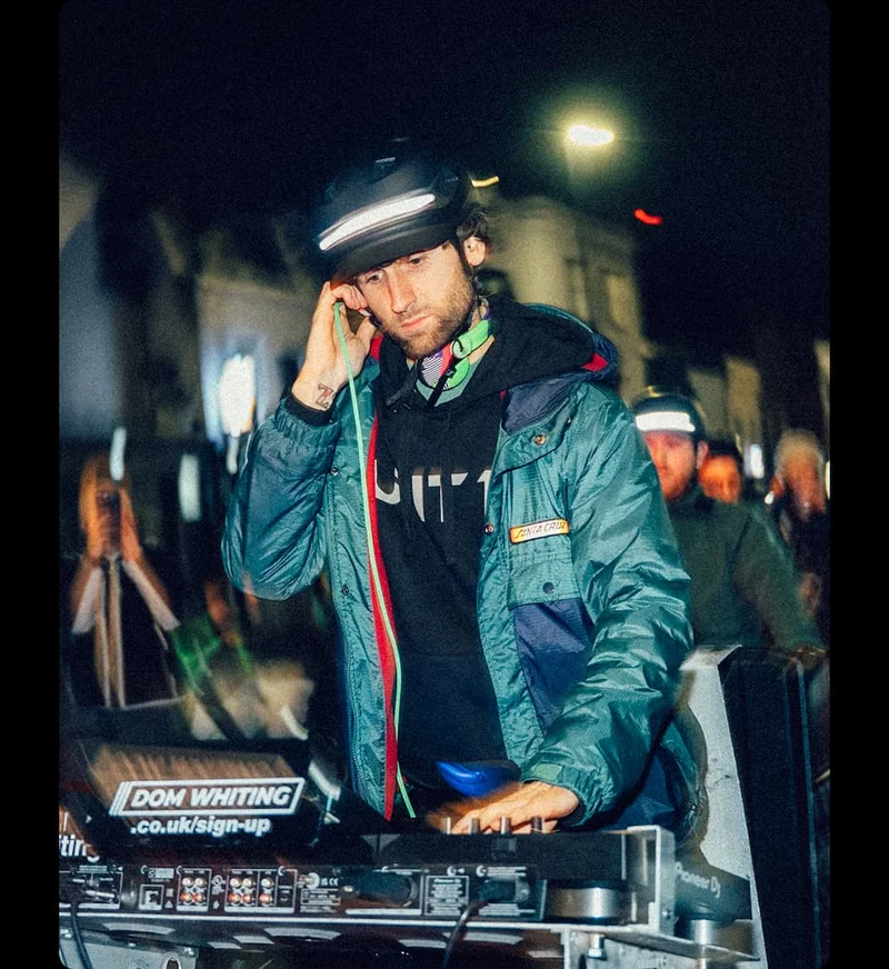 Dom Whiting DJing outdoors at night while wearing an illuminated helmet during a group ride event.