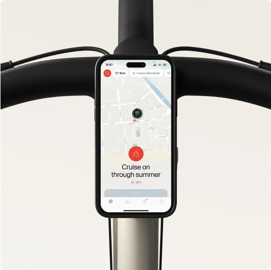 Smartphone mounted on a Cowboy bike’s handlebars, showing the app interface with a map and the text “Cruise on through summer.”