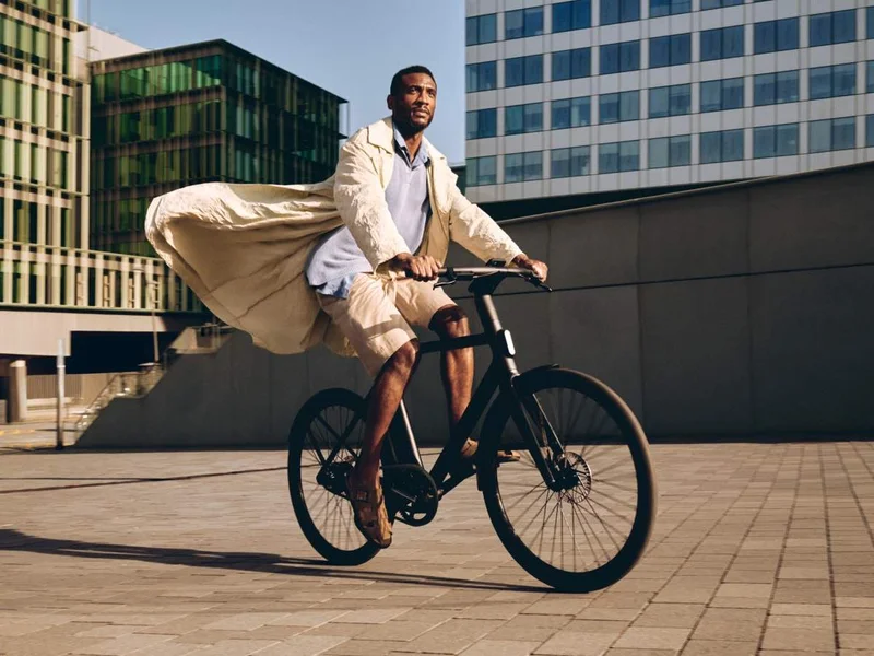 A man riding a black Cowboy bike in an urban setting, wearing a flowing light-coloured coat. The background features modern architecture with glass and concrete buildings.