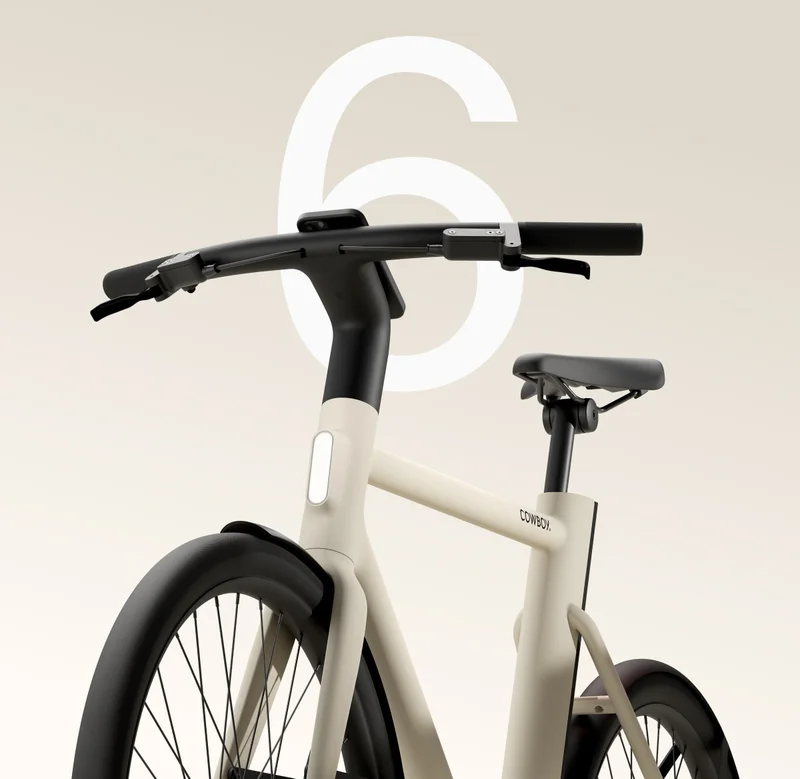 Close-up of the handlebars and front frame of a sand-coloured Cowboy bike, showcasing its minimalist design with an integrated front light. A large white number “6” is visible in the background.