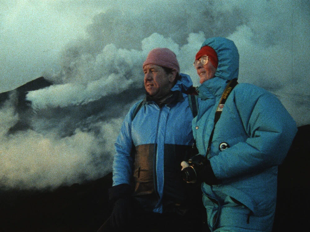 Katia and Maurice Krafft gaze at a volcano in the distance as plumes of ash, steam, and gasses billow behind them.