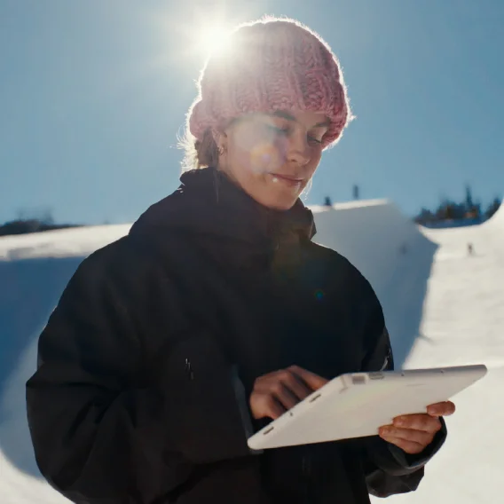 A young woman outdoors in the snow, wearing a black jacket and a pink knitted hat, looking at a white tablet device