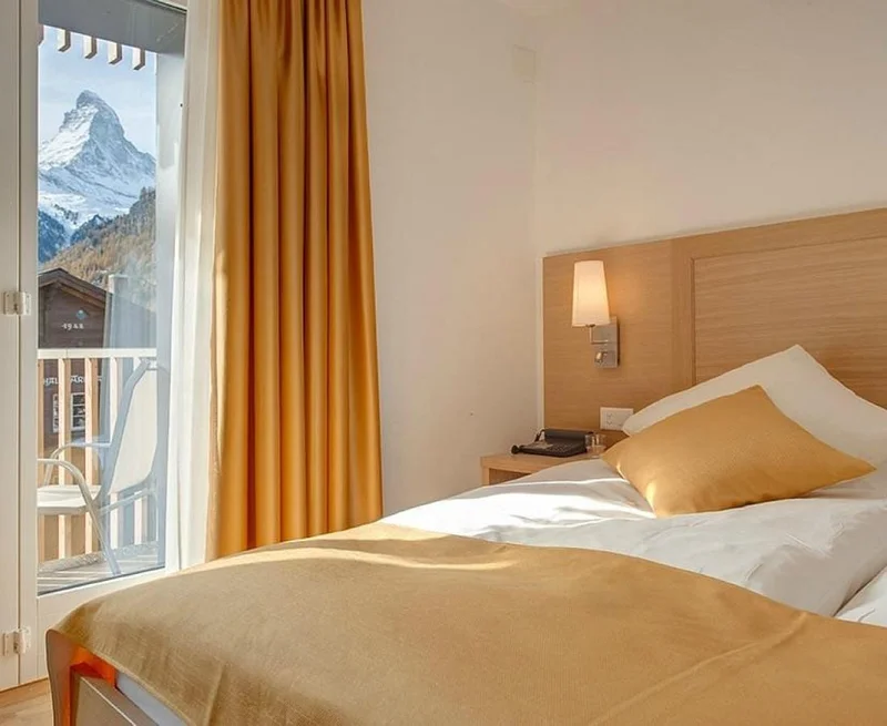 Image description: Bright hotel room with a white bed with a light wood headboard. A glass door leading to a balcony shows a view of a snow-capped mountain in the distance.