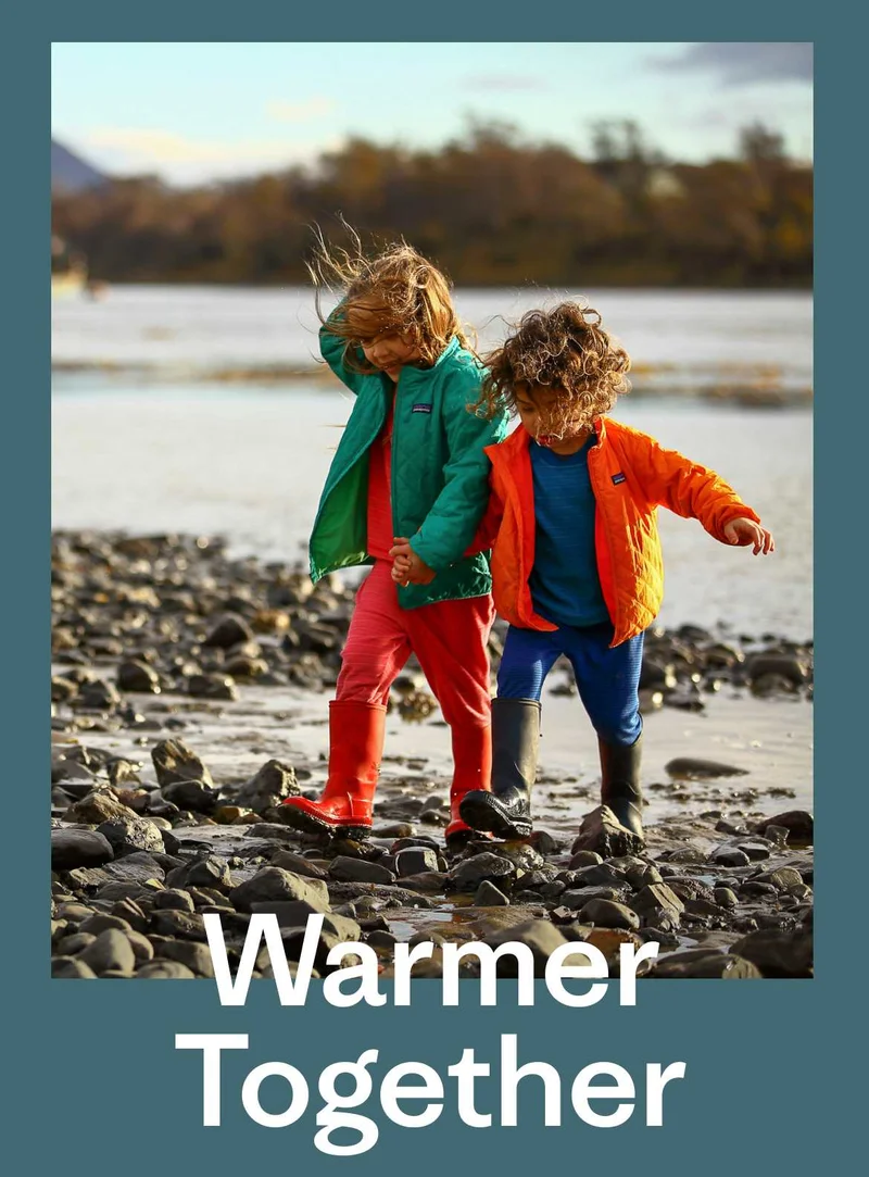 Patagonia. Two little kids walk along the water’s edge in rain boots. Warmer Together.