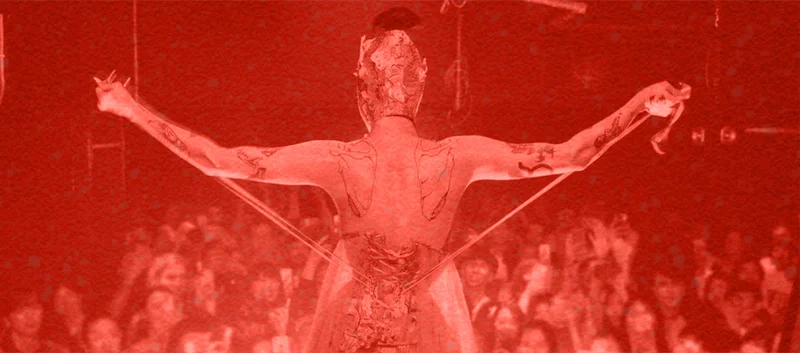 A drag performer in an elaborate costume extends their arms on stage, bathed in red light.