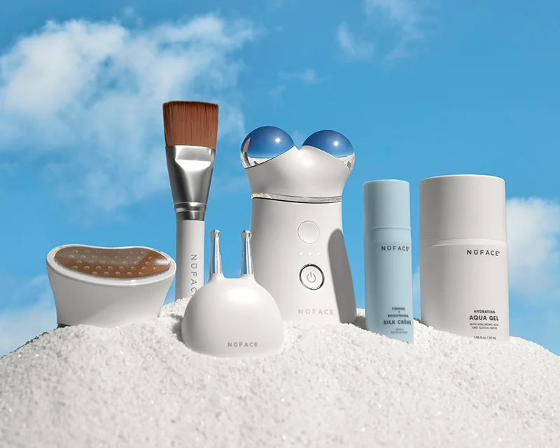 TRINITY+ Complete Set in the sand against a bright sky background. Includes Wrinkle Reducer Attachment, Effective Lip and Eye Attachment, Facial Trainer Attachment, trial-sized Silk Creme Microcurrent Activator and full-sized Aqua Gel Microcurrent Activator