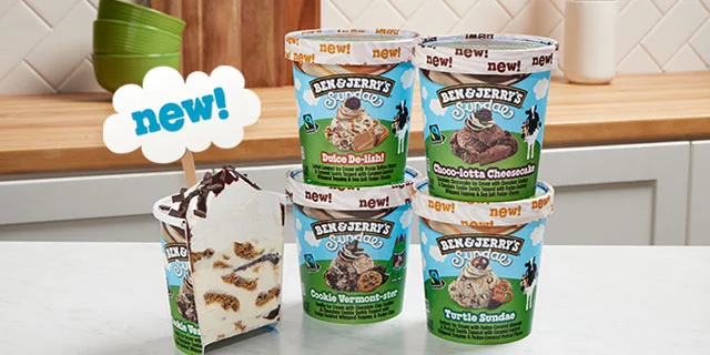 Kitchen setting with four Ben & Jerry's Sundaes pints. One pint is split open in a cross-section, so you can see the layers of ice cream, chunks and swirls, and whipped topping inside.