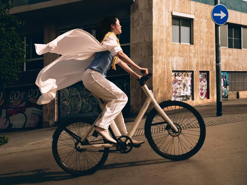 A woman riding a sand-colored Cowboy Cruiser bike through an urban street, her coat billowing behind her. The background features a building with graffiti-covered walls and posters, adding a vibrant city atmosphere.