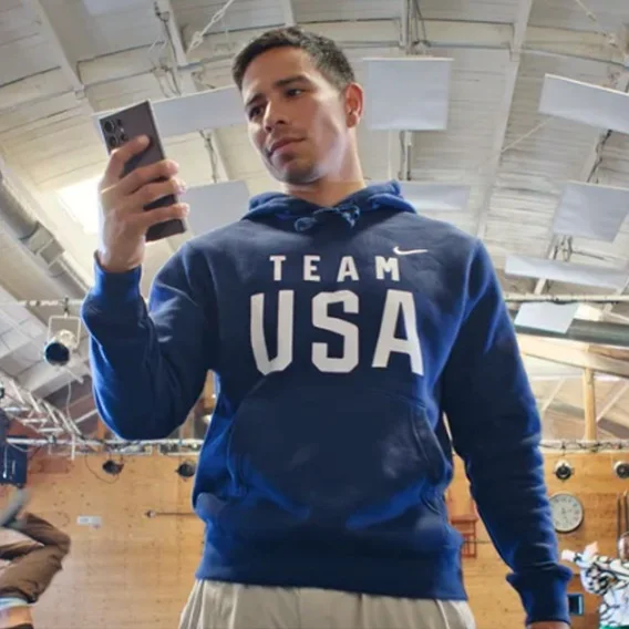A photo of a man wearing a blue “Team USA” sweatshirt holding a phone and looking at the screen. A gym is in the background of the image.