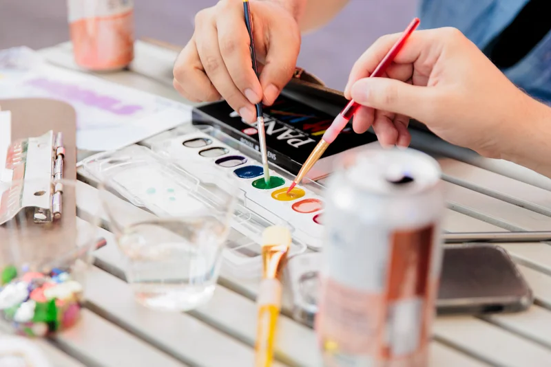 A photograph of two hands dipping paintbrushes in watercolor paint