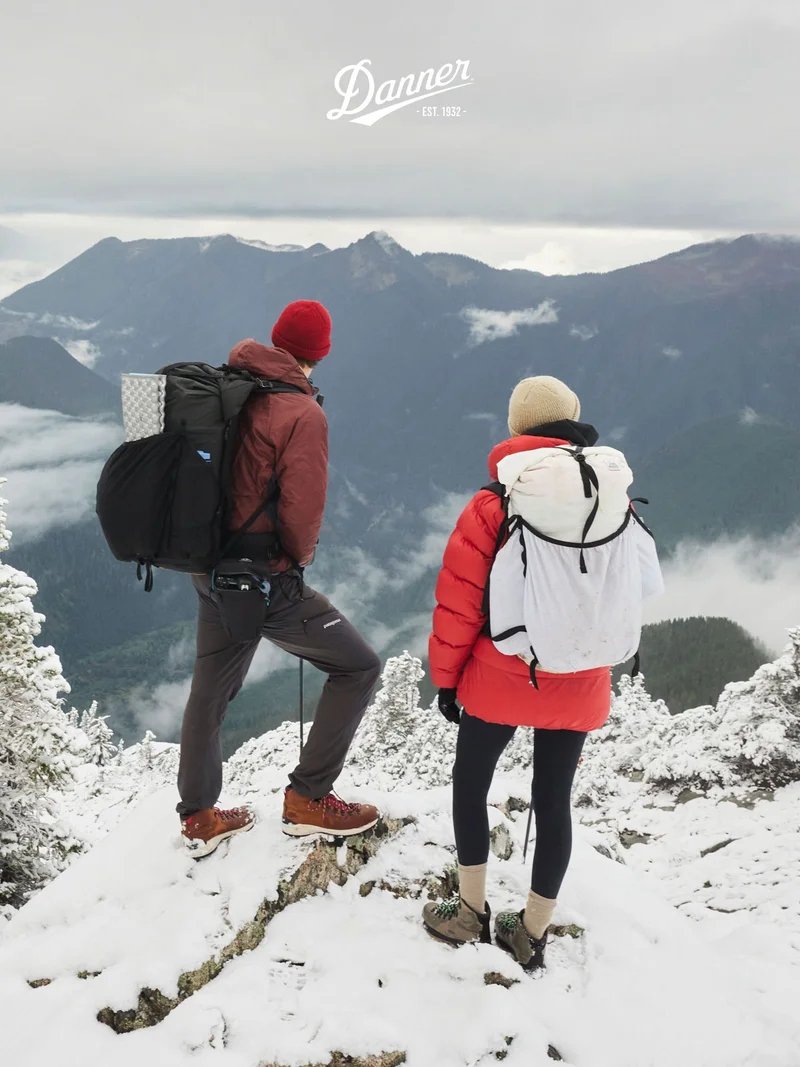 Two hikers, stand on a snowy mountain peak overlooking a range of misty mountains. The man, facing left, wears a red beanie, brown jacket, gray pants, and brown hiking boots with red laces. The woman, facing right, wears a cream beanie, red puffer jacket, black leggings, and Mountain 600 EVO hiking boots. Both carry backpacks and are equipped for cold weather. The Danner logo is displayed at the top.