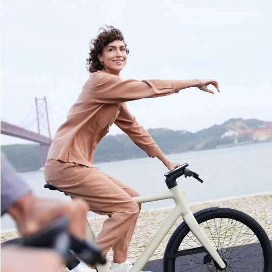 A photograph. A woman rides a Cowboy bike besides a waterway, she's pointing ahead while looking at a comapnion just out of shot beside her.