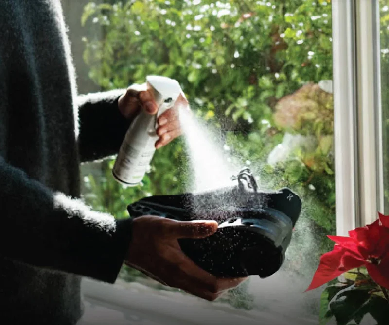 A medium shot shows a person in a dark sweater holding a white spray bottle labeled 'FEET' and spraying a black shoe with a blue accent, with a red po...