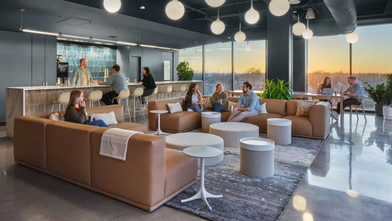 Casual workplace setting with people sitting and standing around a coffee or snack bar talking. A person sitting on a light brown sofa sectional talking on the phone with three people sitting opposite each other, having a conversation.