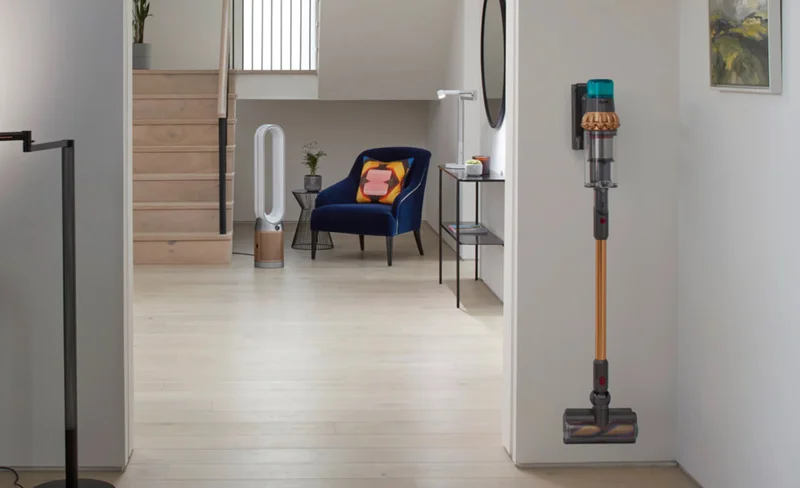 A hallway setting with a Dyson vacuum cleaner in the foreground and a Dyson air purifier in the background, between a staircase and armchair