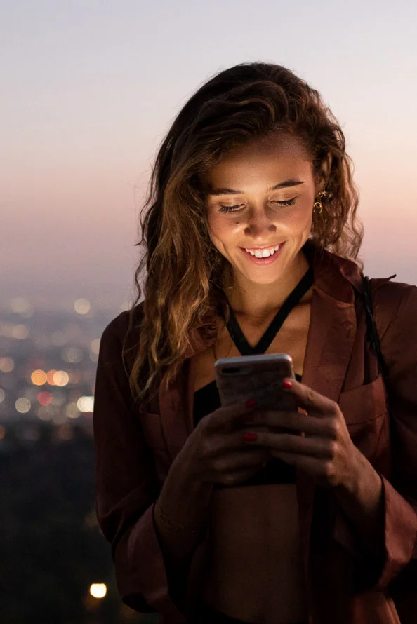 A woman smiles at her smartphone with the city skyline behind her.