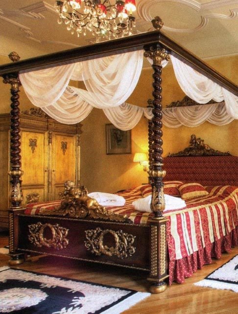 Image Description: The lavish red-and-gold interior of a hotel room in Prague with a chandelier, canopy bed, and a gold wardrobe