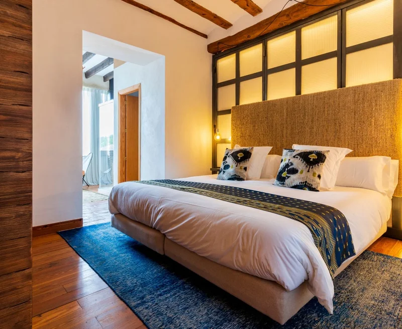 Image description: Hotel room with dark wood accents and a large bed in the center with blue printed pillows and decor.