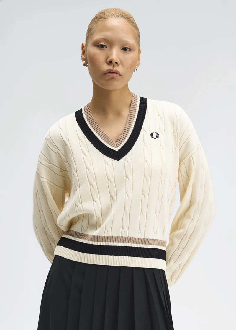 Fred Perry New Releases: female model with slicked back bleached hair stands front-on to the camera with arms behind her back wearing a cream v-neck long-sleeved cable knit jumper with black and mink coloured tipping.