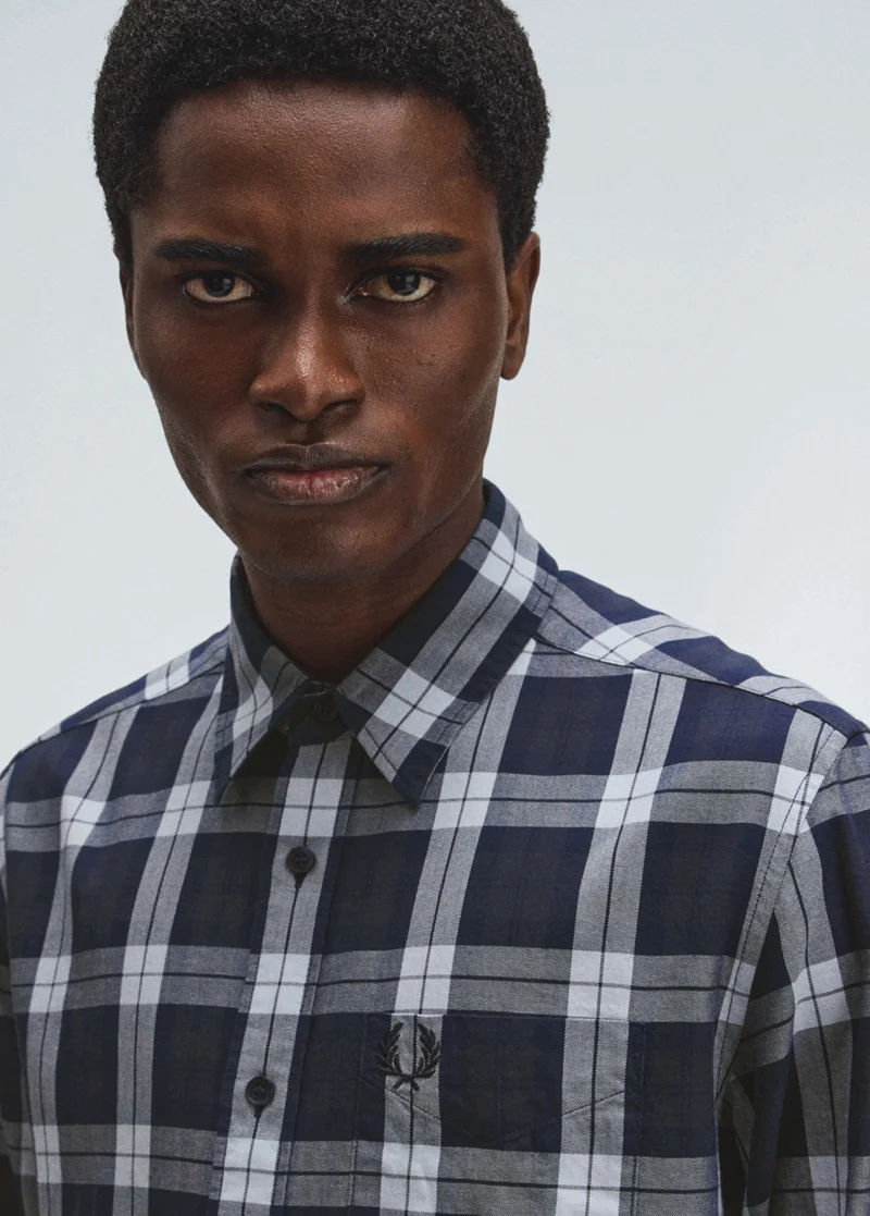Black male model with short hair stares into the camera and is wearing a navy and white checkered button-up collared shirt