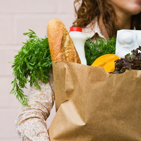 A close-up of a person holding a large paper grocery bag filled with fresh produce, bread, and milk, suggesting a focus on sustainable shopping and healthy eating.