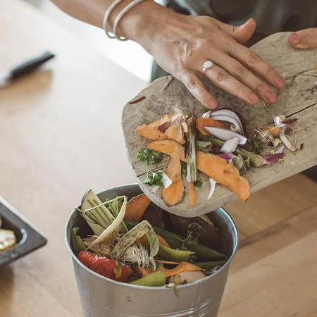 Someone composts vegetable scraps from a cutting board into a bin, with a tray of colorful chopped veggies nearby, highlighting sustainable kitchen habits.