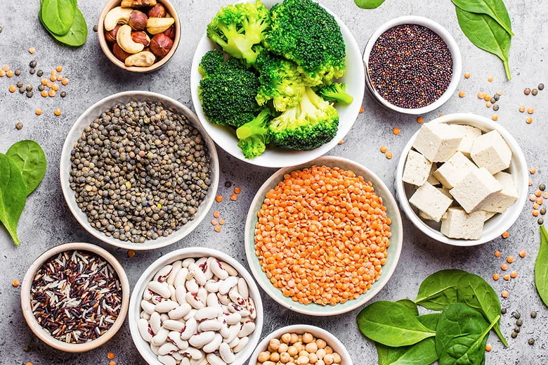 A top-down view of various bowls filled with colorful vegan ingredients, including green and red lentils, white beans, chickpeas, quinoa, wild rice, and broccoli, showcasing a variety of healthy meat alternatives.