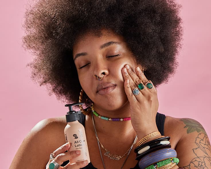 A Black person with an afro and lots of rings and bracelets gently presses lotion into her face.