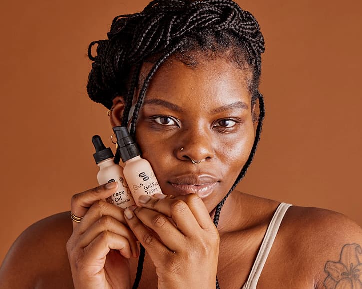 A Black person with box braids up in a crown braid and tendrils coming down with two pink bottles hugged against their cheek.