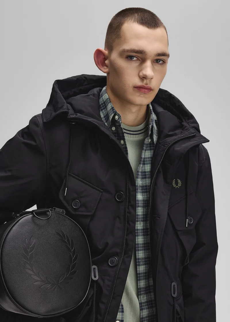A male model wearing a Fred Perry Patch Pocket Puffer Jacket in black holding a black Fred Perry barrel bag