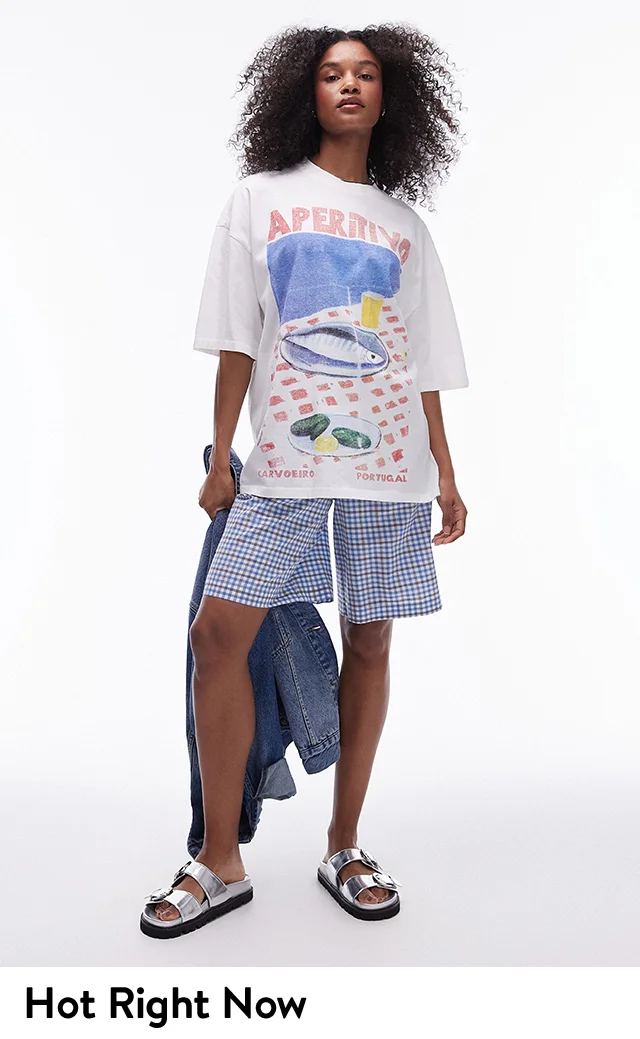 A woman wearing an oversized graphic tee, gingham shorts and silver slide sandals.