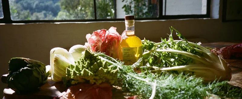 A bottle of west~bourne organic avocado oil surrounded by fresh green produce in a sunlit kitchen.