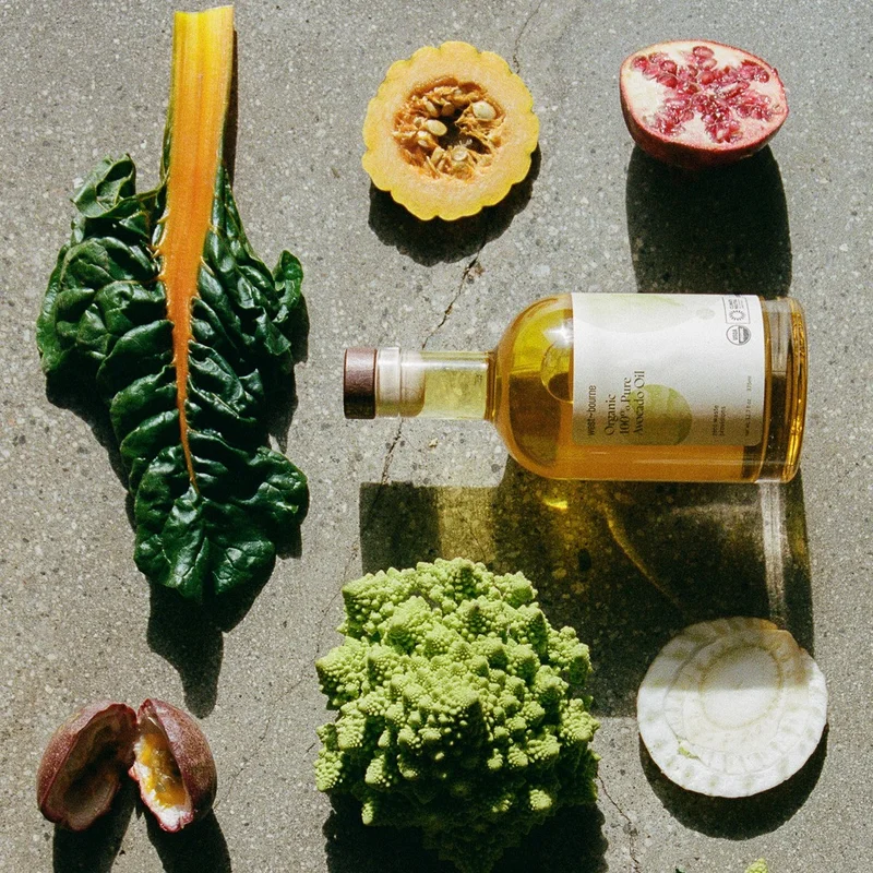 Close-up of a clear glass bottle of west~bourne organic avocado oil surrounded by vibrant heads of pink radicchio, green Romanesco broccoli, and other fresh herbs on a wooden surface.