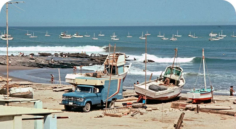 An old photo of a wave breaking near anchored and beached fishing boats.
