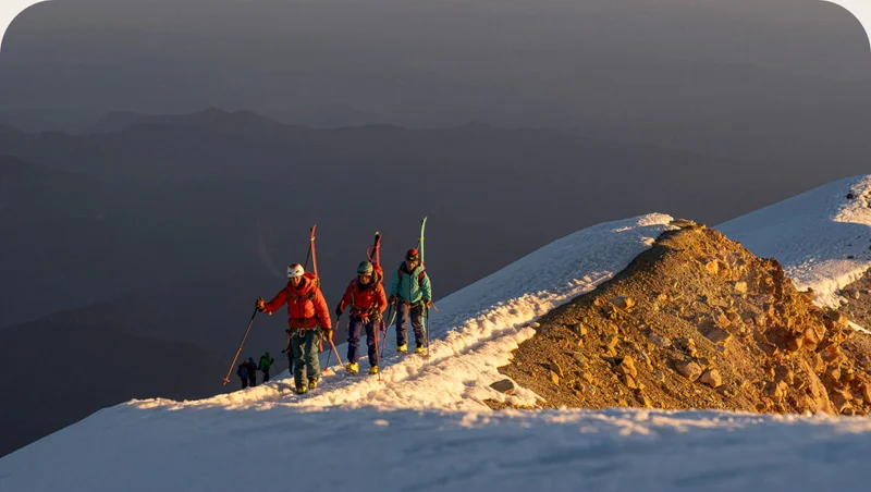 Three backcountry skiers walk along a snowy ridge with their skis on their backs.
