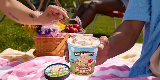 Friends at a picnic, one person holds a pint of Ben & Jerry's Strawberry Cheesecake ice cream while another uses a spoon to eat a scoop.