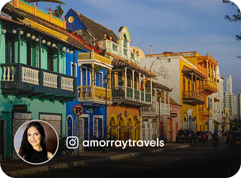 Image Description: Colorful buildings with balconies on a street. A profile photo of a person and the Instagram handle amorraytravels is on the bottom left corner of the image.