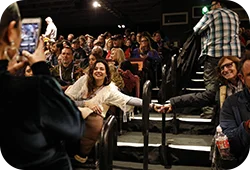 Two women holding hands across an aisle posing for a picture in a crowded theater.
